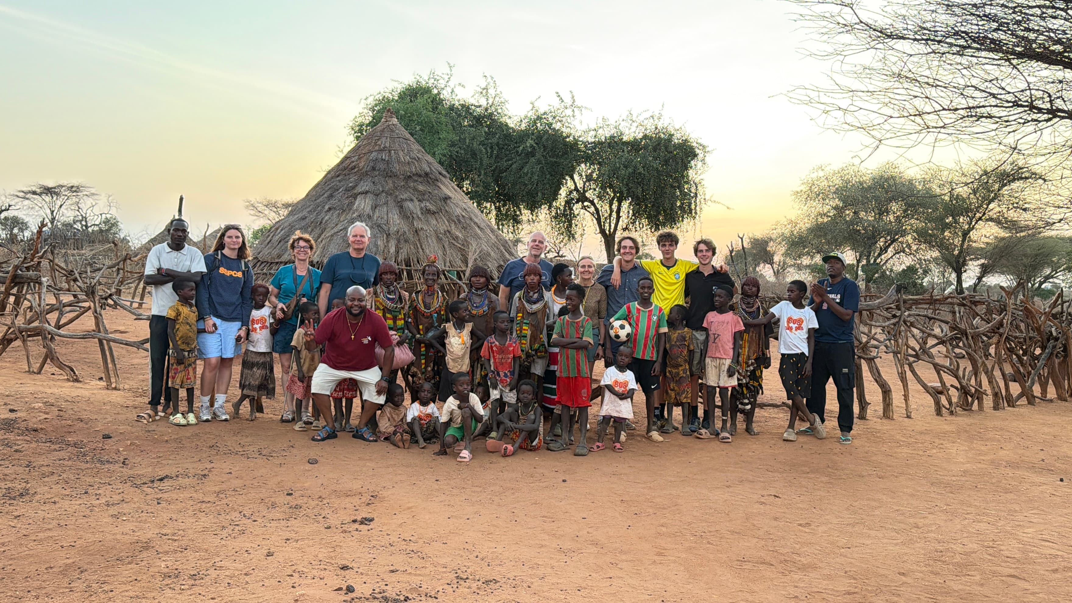A multigenerational family posing with Hamar tribe members and children outside a traditional thatched hut in the Omo Valley