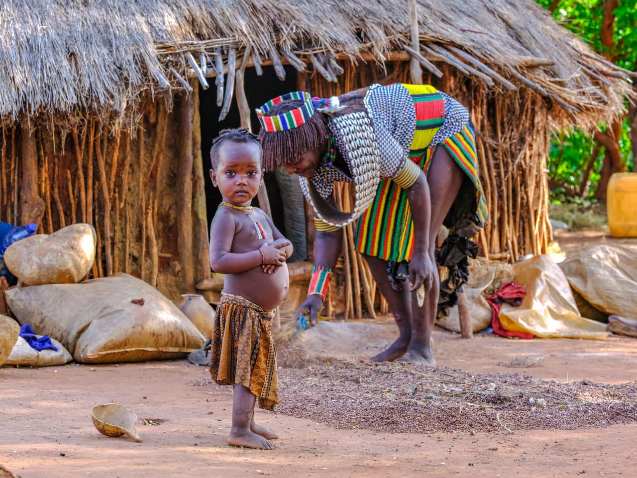 Hamar mother in traditional cowrie shell necklace sweeping grains beside her young child in South Omo