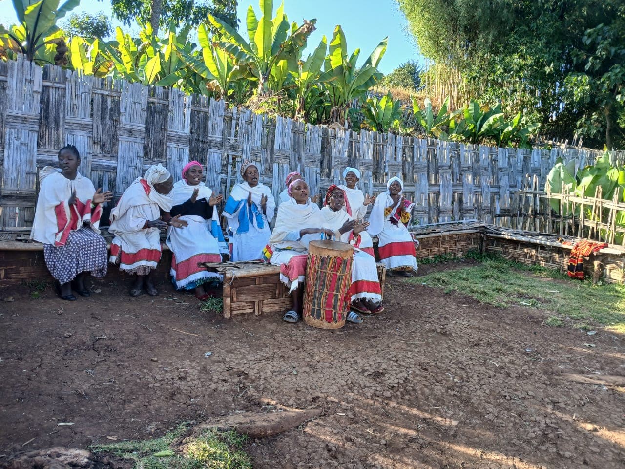Women in traditional dress singing and clapping around a drum during a village ceremony