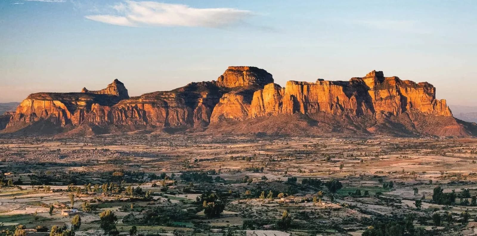 Dramatic sandstone rock formations glowing orange at sunset over the Ethiopian highlands, with green farmland and scattered trees in the valley below